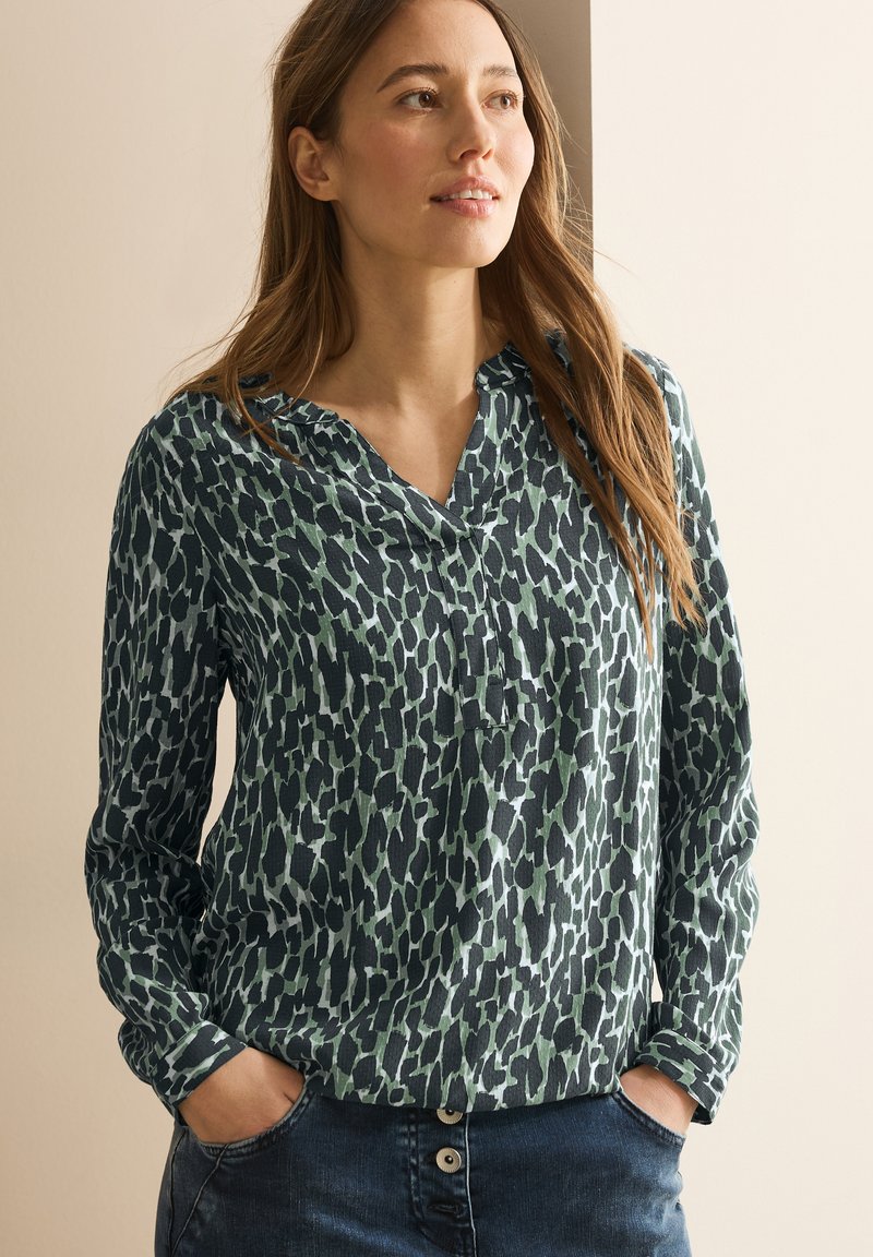 Langsleeve blouse in donker groen met een abstract dierenprint ontwerp, voorzien van een kraag en knopenlijst, gecombineerd met blauwe denim jeans.