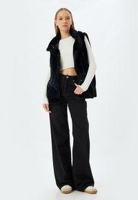 Schwarze Kunstfellweste über einem weißen gerippten Croptop, kombiniert mit hochgeschnittenen schwarzen Hosen und hellen Sneakers.