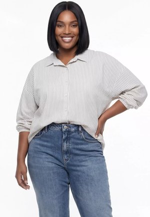 Uśmiechnięta kobieta o czarnych włosach do ramion, ubrana w jasno szarą, paskowaną koszulę na guziki i niebieskie dżinsy, pozująca z jedną ręką na biodrze.