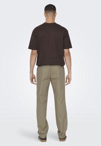 Męski ciemnobrązowy t-shirt z krótkim rękawem, w połączeniu z jasnobeżowymi spodniami. Materiał z mieszanki bawełny. Widok od tyłu, bez widocznych wzorów ani logo.