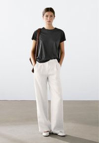 Sort kortærmet t-shirt parret med hvidt vertikalt stribet brede bukser og flade hvide sko, mens der holdes en brun vævet taske.