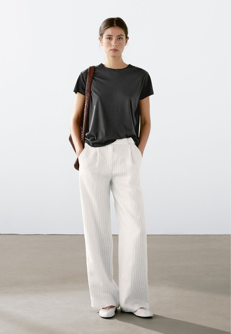 Sort kortærmet t-shirt parret med hvidt vertikalt stribet brede bukser og flade hvide sko, mens der holdes en brun vævet taske.