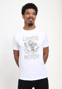Bílý bavlněný tričko s černým grafickým potiskem orla a kytary, s textem "LYNYRD SKYNYRD" v tučném stylizovaném písmu.