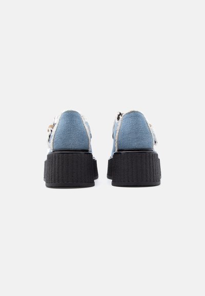 Paire de chaussures à plateforme vue de dos, avec des dessus en denim bleu clair et des semelles épaisses noires texturées sur fond blanc.
