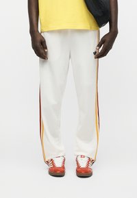 Άνδρας με άσπρο παντελόνι φόρμας Adidas με κίτρινες και κόκκινες ρίγες, κόκκινα αθλητικά παπούτσια Adidas, κίτρινο πουκάμισο και μαύρη τσάντα ώμου.