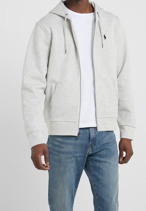 Mand iført lysegrå lynlåstrøje med forlommer, hvid t-shirt og blå jeans foran ensfarvet baggrund.