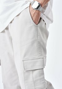 Pantaloni cargo grigi chiari con tasche laterali, realizzati in tessuto resistente. Presentano dettagli cuciti e una vestibilità rilassata. Orologio argento al polso.