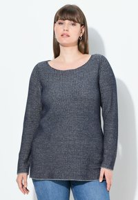 Žena s rovnými hnědými vlasy, která má na sobě kruhové náušnice, tmavě modrý a bílý pruhovaný svetr s dlouhým rukávem a modré džíny, stojí proti bílé zdi.