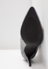 Chaussure à talon haut à bout pointu avec semelle noire et dessus en tissu à carreaux gris, vue du dessous, reposant contre un coin blanc.