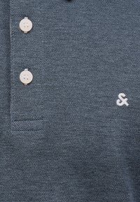 Κοντινό πλάνο μπλε πικέ πόλο πουκάμισου με δύο λευκά κουμπιά και ένα μικρό λευκό κεντημένο σύμβολο αμφικέ στον θώρακα.