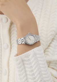 Sudraba metāla pulkstenis ar oktagona formas ciparnīcu, baltu pērlīšu dialu, rozā zelta stundu rādītājiem un kristāla akcents, valkāts uz rokas.