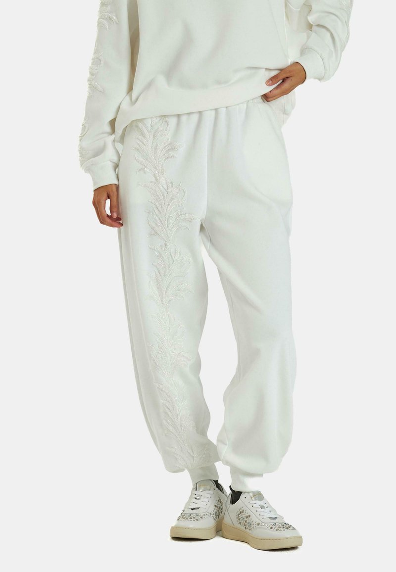 Pantalones de chándal blancos con patrones de hojas texturizados y bordados en los laterales. Con un ajuste relajado y cinturilla elástica. Combinados con zapatillas decorativas.