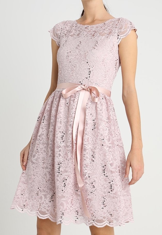 Robe en dentelle lavande avec manches courtes, paillettes et ceinture en ruban de satin nouée en un nœud à la taille, portée par une femme debout.