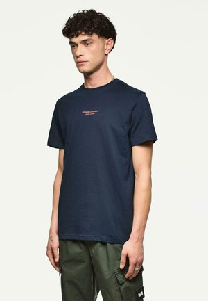 Man met krullend haar in een marineblauw T-shirt met de tekst "WEEKEND OFFENDER ADMIT NOTHING" en groene cargobroek, staande tegen een effen achtergrond.