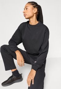 Μαύρο φούτερ και παντελόνι από μαλακό ύφασμα, με μικρό λογότυπο στο στήθος και στο πόδι. Συνδυασμένα με μαύρα αθλητικά παπούτσια και κάλτσες.