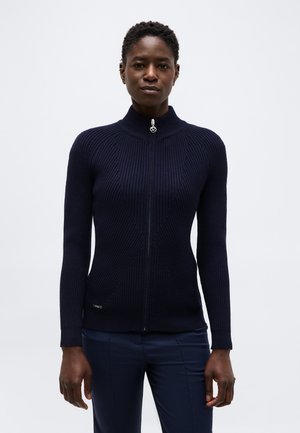 Femme portant un pull zippé côtelé bleu marine ajusté et un pantalon bleu marine assorti, debout devant un fond clair uni.
