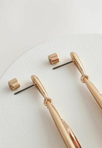 Guldfarvede metaløreringe med et forlænget, buet design og en cylindrisk studs. Teksturoverflade og poleret finish.