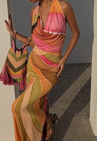 Femme en robe dos-nu colorée à motifs, tenant un sac tissé multicolore, debout à côté d'un mur clair projetant des ombres.