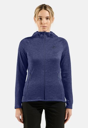 Woman wearing a blue zip-up hooded jacket with zippered side pockets and black pants, standing against a plain light background.