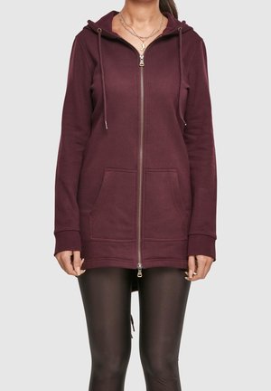 Kvinde iført en lynlåst vinrød hættetrøje med forlommer og sorte leggings, vist fra nakke til midt på låret mod en neutral baggrund.