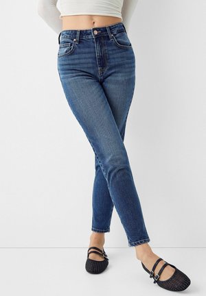 Femme portant un jean skinny bleu taille haute et des ballerines noires tissées, debout les jambes croisées sur un fond blanc.