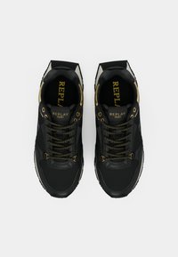 Une paire de baskets noires Replay avec des accents dorés, des lacets noirs avec des points jaunes, et "REPLAY 1981" écrit sur la languette et les semelles intérieures.