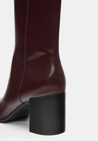 Burgundowe skórzane buty za kolano o błyszczącej powierzchni. Posiadają masywny czarny obcas i widoczne szwy wzdłuż boku.