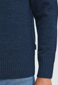 Mørkeblå strikket genser er slitt over lyseblå denimjeans på en persons arm og liv.