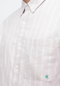 Lys rosa skjorte med knapper, hvite vertikale striper, en lomme på venstre side og en grønn logo sydd på lommen.