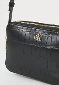 Svart präglad lädercrossbodyväska med guldfärgad dragkedja, logotyp i guld och texturerat krokodilmönster på utsidan.