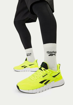Gambe con leggings neri, calzini bianchi Reebok e scarpe da corsa Reebok giallo brillante con suole bianche su uno sfondo uniforme.