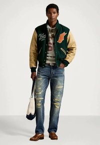 Veste de varsity verte et beige avec des détails brodés, sweat-shirt gris, jeans bleus déchirés, mocassins marron et un sac à bandoulière.