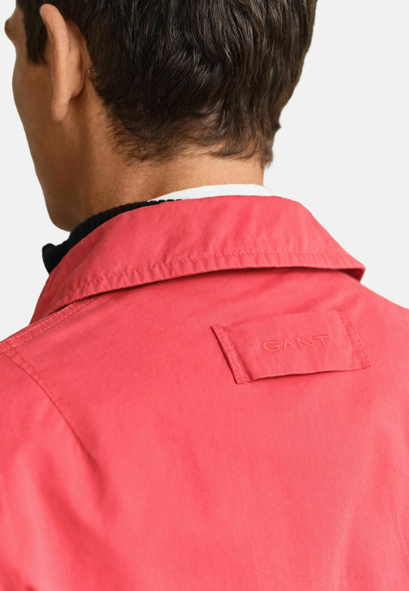 Homme portant une veste rose corail avec un patch logo GANT visible sur le haut du dos, tourné de dos à la caméra.