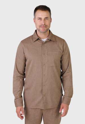 Mand iført en brun langærmet skjorte med knapper og en frontlomme, stående mod en ensartet lysegrå baggrund.