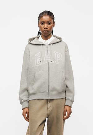 Fiatal személy fonott hajjal, világosszürke, cipzáras kapucnis pulóvert visel, amelyen a "GAP" felirat olvasható, és bézs színű nadrágot, sima háttér előtt állva.