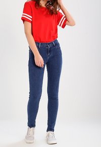 Camisa corta roja de manga corta con rayas blancas, combinada con jeans ajustados azules y zapatillas gris claro. Enfocado en un estilo casual y un buen ajuste.