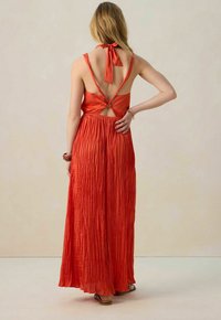 Robe maxi en tissu rouge vif, avec un col halter, un dos nu et une texture plissée. Longueur longue avec une taille ajustée et une jupe fluide.