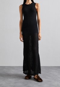 Robe noire sans manches, longueur genou, en tissu tricoté ajouré, avec un col rond et un motif texturé. Associée à des chaussons doublés de fourrure.