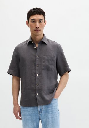 Mann trägt ein kurzes Hemd in Kohlefärbung mit Brusttasche und hellblaue Jeans, steht vor einem schlichten weißen Hintergrund.