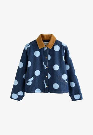 Mujer rubia con chaqueta y pantalones de mezclilla azul oscuro con grandes lunares azul claro, de pie en interiores con la mano en el bolsillo.