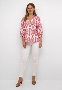Rote und weiße gemusterte Bluse mit Puffärmel und V-Ausschnitt, kombiniert mit weißen Skinny-Jeans und braunen Absatzsandalen. Glatte Stoffoberfläche.