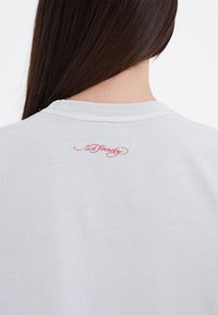 Lysegrå bomulds t-shirt med rund hals, med et rødt broderet logo på den øverste ryg. Glat tekstur, enkelt design.