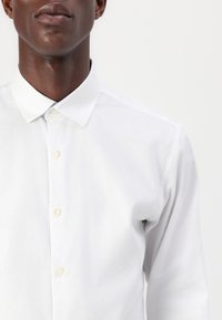 Biała koszula na guziki z klasycznym kołnierzykiem, z delikatną fakturą i jasnoniebieskim akcentem wewnątrz kołnierzyka. Widać pięć guzików.