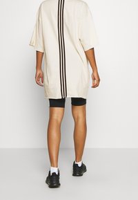 Camiseta de gran tamaño color crema con mangas cortas, que presenta dos rayas verticales marrón oscuro en el centro, combinada con pantalones cortos de ciclismo negros y zapatillas negras.