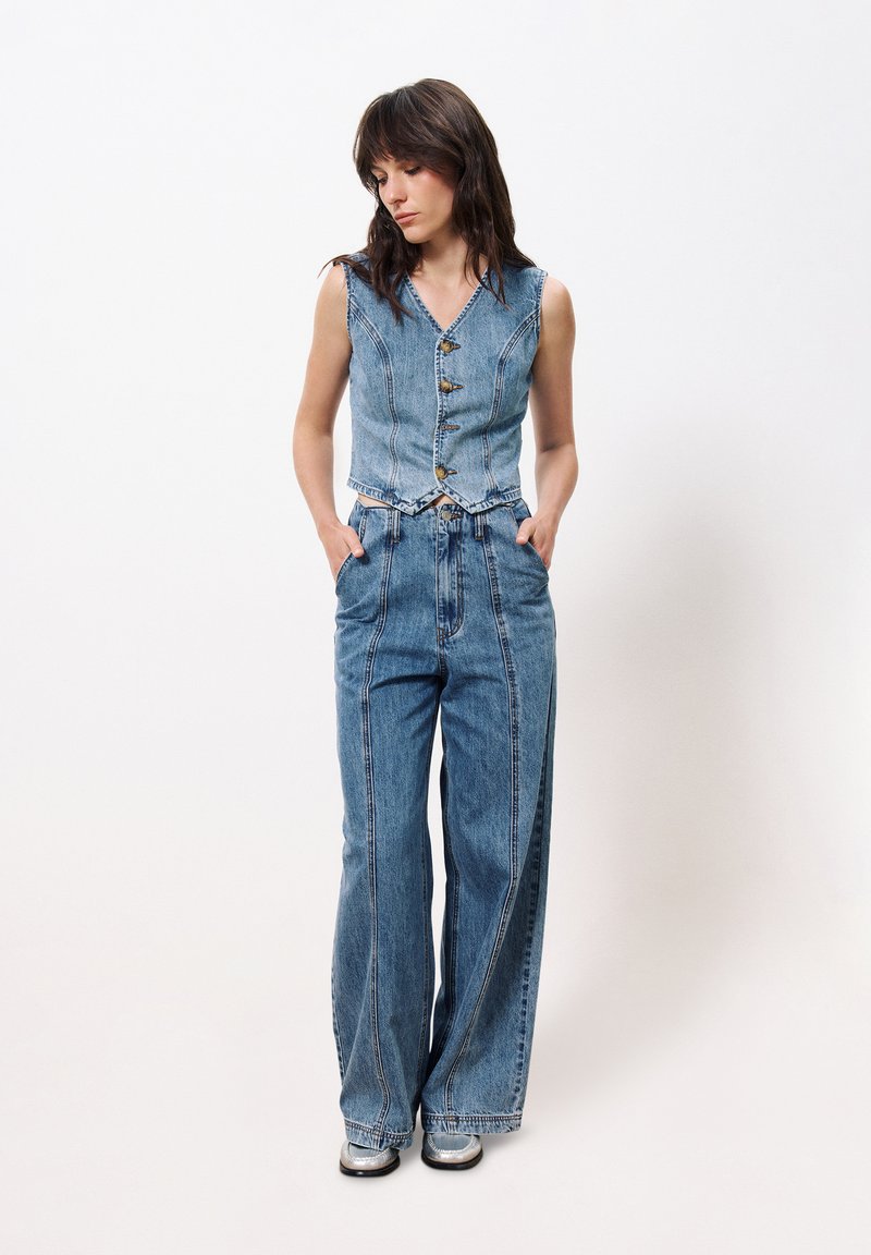 Gilet en denim avec un col à revers et une fermeture à boutons sur le devant, associé à un jean à jambe large avec des coutures verticales et une coupe décontractée.