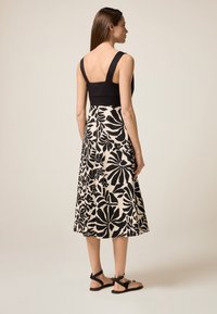 Schwarz-ivorfarbenes Kleid mit Blumenmuster, breiten Trägern, tailliertem Oberteil und fließendem Rock. Das Model trägt flache schwarze Sandalen.