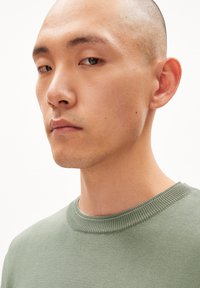 Suéter de punto en verde claro con cuello redondo texturizado de canalé. Modelo mostrado con una expresión neutral y cabeza afeitada, mirando levemente hacia la izquierda.