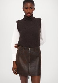 Maglione a collo alto in lana marrone scuro sovrapposto a una camicia bianca, abbinato a una mini gonna in pelle marrone con zip frontale e quattro tasche.