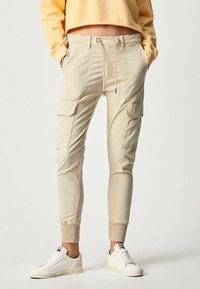 Beiga cargobyxor med elastiska ankle cuffs, dragsko i midjan och sidofickor. Kombineras med vita sneakers. Slätt, lättviktigt tyg.