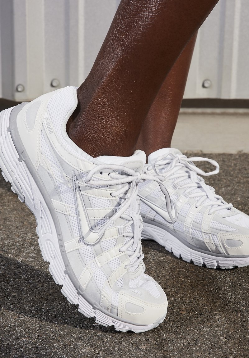 Personne à la peau foncée portant des chaussures de running Nike blanches avec des empiècements en mesh et synthétiques sur l'asphalte près d'un mur clair.